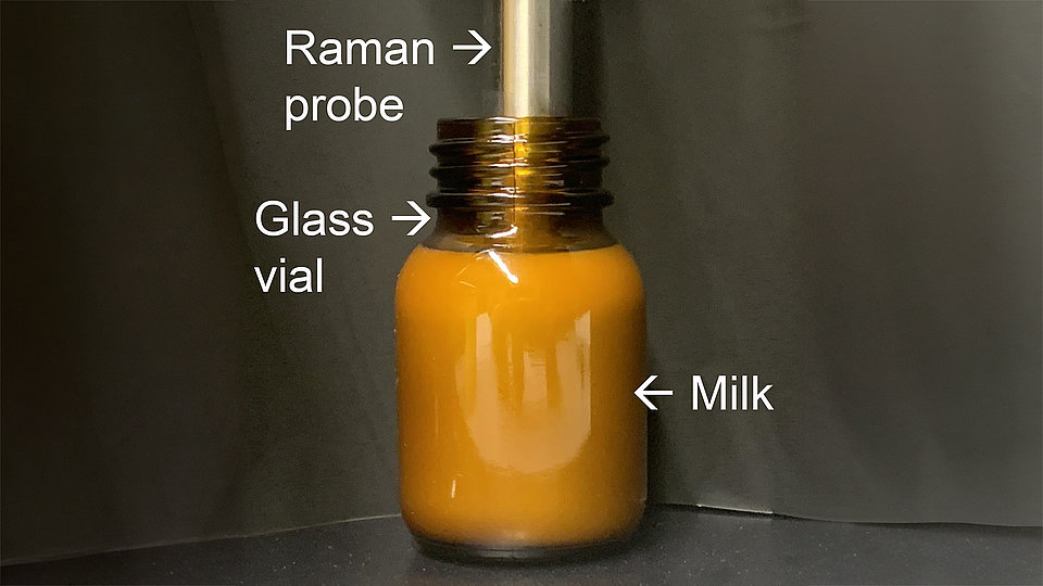 Close-up photo of a metal Raman probe inside a brown glass vial. Labels are used to identify the key components: "Raman probe" points to the top part of the metal probe, "Glass vial" points to the neck of the transparent container, and "Milk" points to the liquid filling the vial.