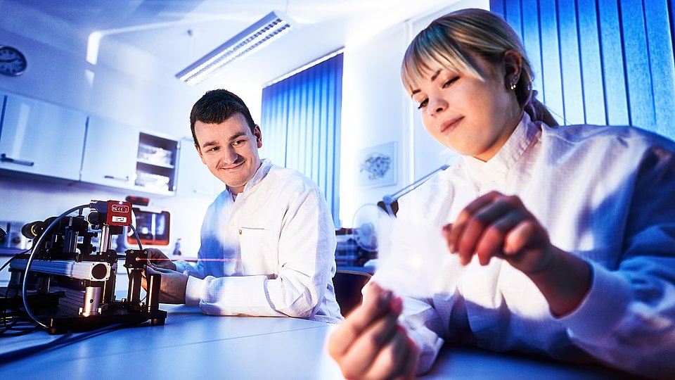 Two people in a lab wearing white coats are working with a diode laser. A woman is holding a small component, while a man looks in her direction and smiles. The scene depicts a modern laboratory with technical apparatus.