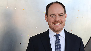 Portrait photo of a middle-aged man in a suit and tie, smiling in front of a bright, minimalist wall. He has short brown hair and a neatly trimmed beard.