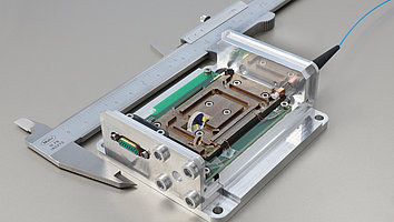 The photo shows a laser module next to a measuring caliper, which illustrates that the module is no larger than 10 cm. Various lenses and a crystal can be seen in the module. The module is coupled with a fiber that guides the pump radiation into the module.