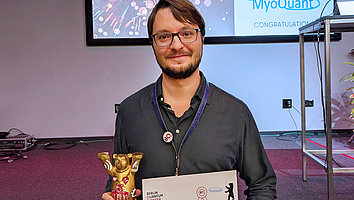 Preisträger des Jury-Preises des Berlin Quantum Pioneer Inkubationsprogramms, Sascha Neinert, steht vor einem Bildschirm mit dem Logo der Veranstaltung. Er hält einen goldenen Berliner Bären im Miniaturformat und die Urkunde in der Hand. Er strahlt in die Kamera.