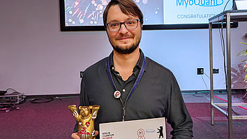 Winner of the Berlin Quantum Pioneer incubation program jury prize, Sascha Neinert, stands in front of a screen with the event's logo. He is holding a miniature golden Berlin Bear and the certificate in his hand. He smiles into the camera.