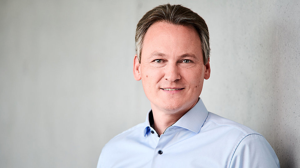 The photo shows Patrick Scheele, Scientific Managing Director of the FBH, standing in front of a gray wall in a light-colored shirt.