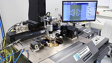 A semiconductor measurement setup with probes positioned on a wafer and a computer screen displaying circuit patterns.