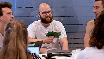 Four students sitting at a table examining a circuit board that comes from the inside of a router. One student is holding the circuit board in his hand, the other participants are discussing it.