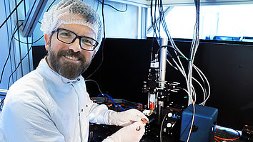 Dr. Paul Crump in the laser laboratory at the Ferdinand-Braun-Institut. He is wearing laboratory clothing with a lab coat and hood and smiling at the camera, holding a diode laser in his hand. In the background you can see the laboratory where the power and efficiency limits of diode lasers based on gallium arsenide are investigated. 