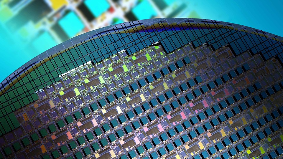 Nahaufnahme eines Halbleiter-Wafers mit zahlreichen Mikrochips, die in einem Gittermuster angeordnet sind und aufgrund der Lichtbeugung verschiedene Farben reflektieren, vor einem unscharfen, bunten Hintergrund.