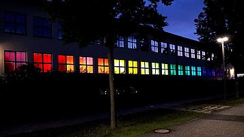 The photo shows the outside of the Ferdinand-Braun-Institut at night with colorfully illuminated windows.