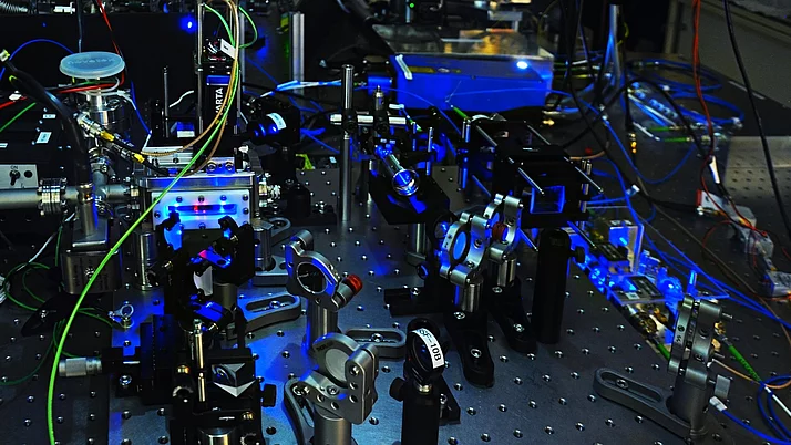 Experimentelle Apparatur mit verschiedenen optischen Komponenten, Metallstützen und bunten Kabeln. Die Struktur wird von blauen und grünen Lichtern beleuchtet.