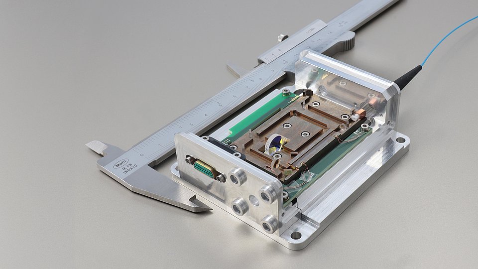 Close-up of a micro-module for quantum interference, mounted in a holder and placed next to a caliper for size reference. The module features delicate electronic and optical components for precise applications.