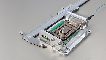 Close-up of a micro-module for quantum interference, mounted in a holder and placed next to a caliper for size reference. The module features delicate electronic and optical components for precise applications.