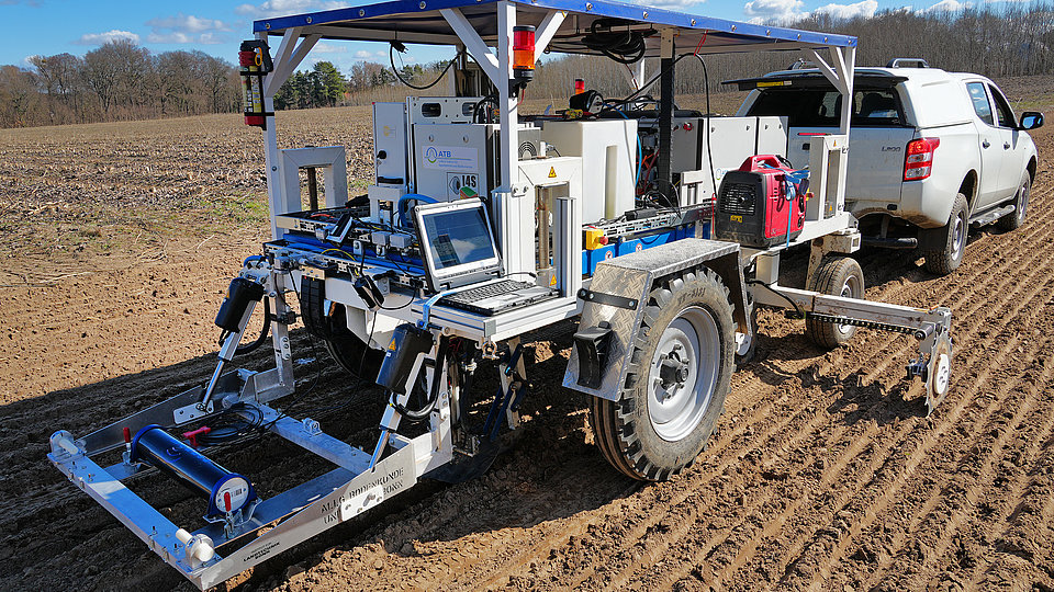 Mobile soil mapping with Raman spectroscopy An agricultural vehicle with a sensor system is standing on a trailer in a field. It has large tires, a platform with a laptop and various technical components. A pickup truck can be seen in the background pulling the trailer. The sky is clear with some clouds.