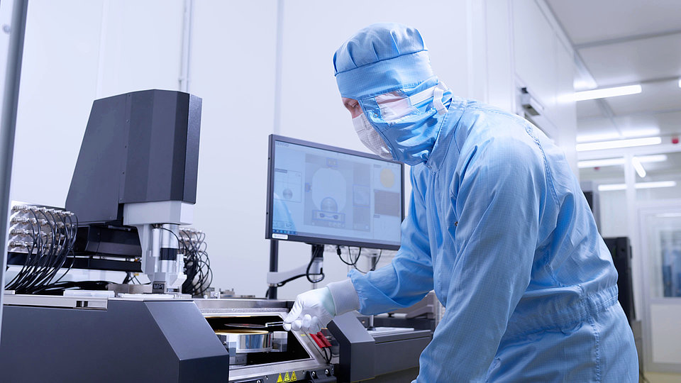 Ein Techniker in Reinraumkleidung bedient moderne Halbleiterfertigungsgeräte. Im Hintergrund ist ein Labor mit einem Monitor zu sehen, der waferbezogene Daten anzeigt.