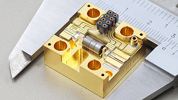 The image shows a diode laser, known as a Master Oscillator Power Amplifier (MOPA), with a caliper as a size reference. The device consists of a gold-colored, rectangular metal block with precise holes, recesses, and, in the front right section, a diode laser that is electrically connected with wires. Additional components, such as lenses, are distributed across the block, which is surrounded by fine conductor paths and structures. A dark gray, round-shaped optical isolator is located in the middle of the device. The caliper scale in the background indicates the small dimensions of the device.